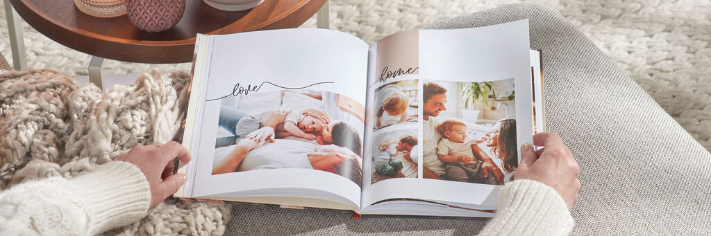 Zwei Hände blättern in einem aufgeschlagenen CEWE FOTOBUCH. Darin sind mehrere Fotos einer Familie sowie die Worte „Love“ und „Home“ zu sehen. Das Fotobuch liegt auf einem grauen Sofa. Links daneben steht ein Tisch mit Teelichtern und einer Pflanze.