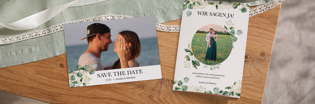 Auf einem Holztisch mit hellgrüner Tischdecke und weißen Bändern liegen zwei Grußkarten im ähnlichen Design. Die linke Postkarte ist eine Save the Date-Karte, die Klappkarte rechts ist eine Hochzeitseinladung.