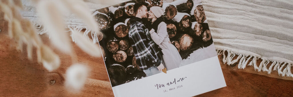 Ein CEWE FOTOBUCH liegt auf einem Tisch. Das Cover zeigt ein Foto der Bloggerin Youdiful mit ihrem Freund Lukas sowie ihr selbst gestaltetes Clipart "you and me" und ein Datum.